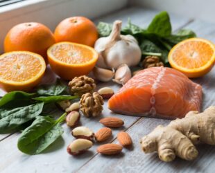 Uma mesa com alimentos coloridos: 🍊 Laranja 🧄 Alho 🫚 Gengibre 🥬 Espinafre 🥜 Castanhas 🐟 Salmão