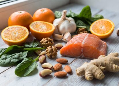 Uma mesa com alimentos coloridos: 🍊 Laranja 🧄 Alho 🫚 Gengibre 🥬 Espinafre 🥜 Castanhas 🐟 Salmão